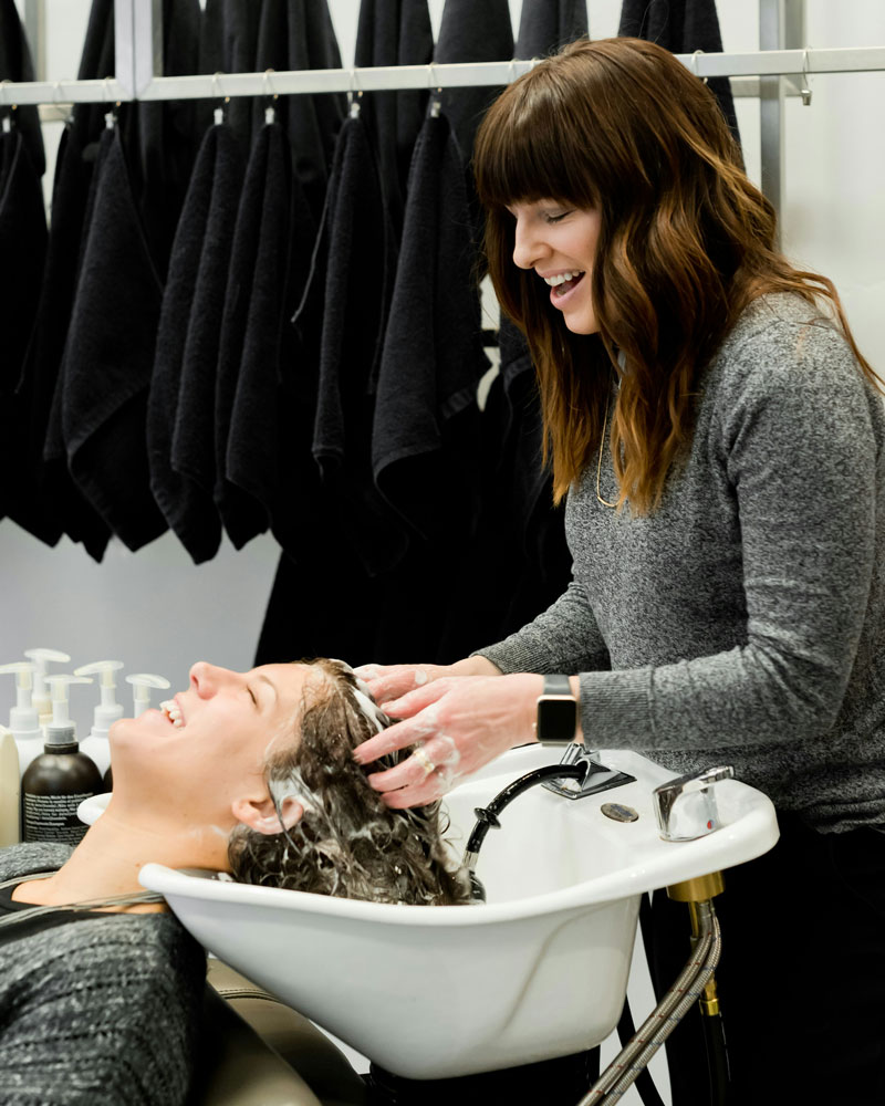 hair stylist washing client's hair at shampoo bowl in salon.