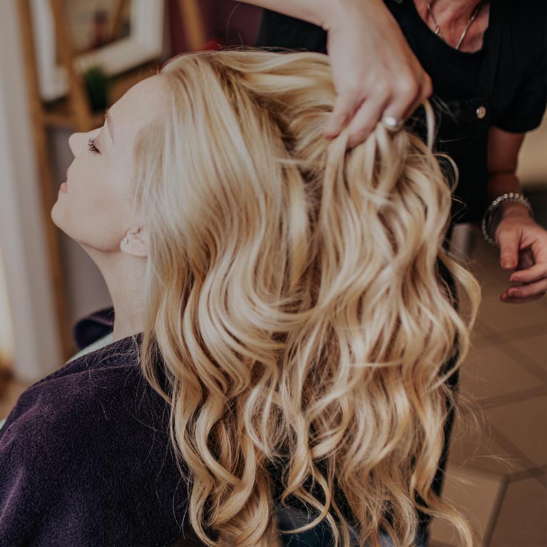 Hairstylist running fingers through a woman's long, curly blonde hair.